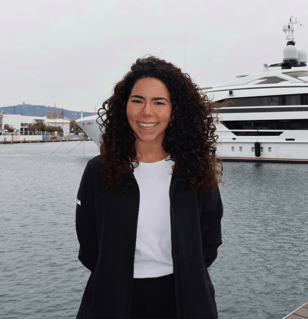 Kiara Arenas smiles while standing near a large docked yacht on a cloudy day.